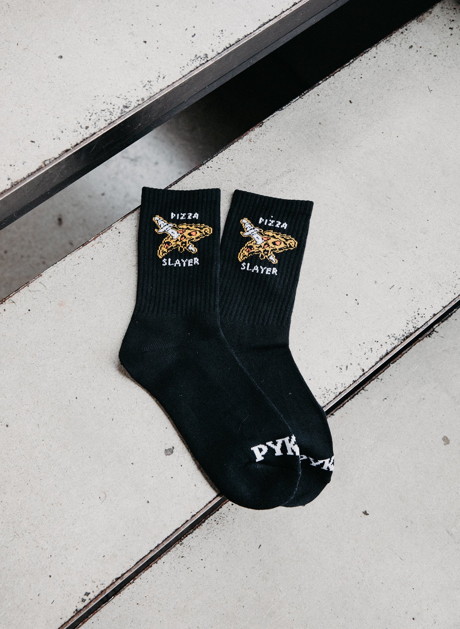 Close-up of Pyknic Pizza Slayer Socks, featuring snarky 'Pizza Slayer' knit design on Black cotton-polyester crew socks.